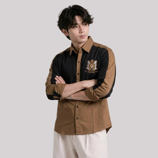 A man modeling a long-sleeve button-up shirt with a color-blocked design featuring black on the right side and brown on the left side and sleeves. The shirt has a brown collar, a brown embroidered crest on the left chest, and is paired with off-white trousers against a solid gray studio background.
